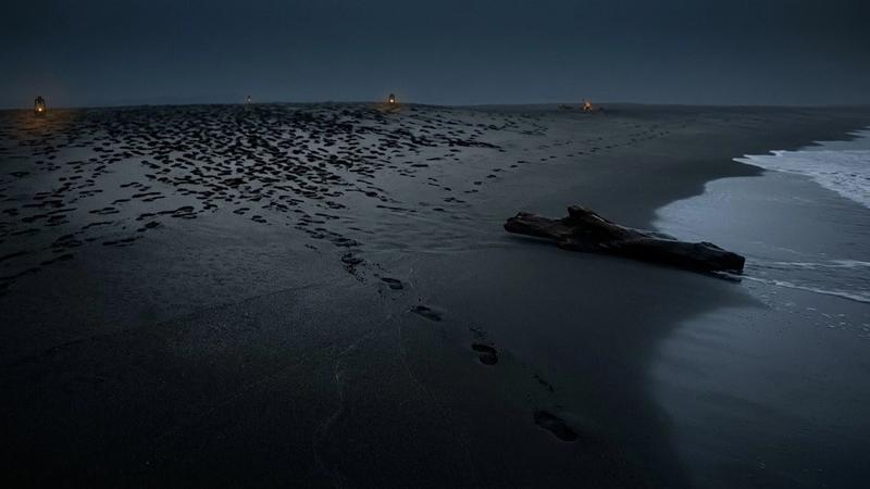 Dark beach at night with scattered footprints and washed-up driftwood. Lanterns glow in the distance, creating a mysterious mood.
