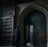 A person stands before a large wooden doorway inside a dimly lit library, surrounded by towering bookshelves and illuminated by light streaming through an arched window.