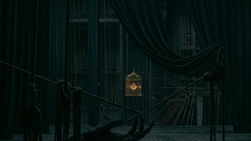 A dimly lit theater stage draped in dark curtains, with rigging ropes and a single illuminated birdcage hanging at the center, creating a dramatic and mysterious atmosphere.