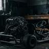 Bare go-kart chassis with an exposed engine in a dimly lit workshop, illustrating a modular mechanical foundation before the bodywork is added.