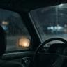 View from inside a parked car on a rainy night, with raindrops covering the windows and blurred streetlights glowing in the distance.