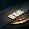 A minimalist, cinematic hero image in a moody, dark tone for a blog post. A smartphone lies flat on a dark, textured surface that mimics continuous, elegant flowing waves. The phone screen displays an interface of the Amazon mobile app with product listings. In the foreground, a prominent, metallic bronze Amazon smile logo rests naturally alongside the phone, catching soft, warm diffused lighting amidst heavy shadows and a desaturated charcoal background.