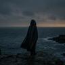 A cloaked figure stands alone on a rocky coastline overlooking a dark, stormy sea beneath heavy clouds, creating a moody and atmospheric scene.