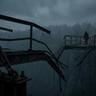 A moody, desaturated photograph shows a person carrying a small lantern across a narrow, rickety suspension bridge over a dark, deep chasm in a misty forest. The near side of the bridge is made of rusty metal and heavy chains, with the path abruptly broken and collapsed into the abyss. The hooded figure walks on the remaining part of the bridge towards a faint, distant light on the far, wooded bank. The scene has soft, diffused light and a grainy, cinematic, and melancholic editorial feel.