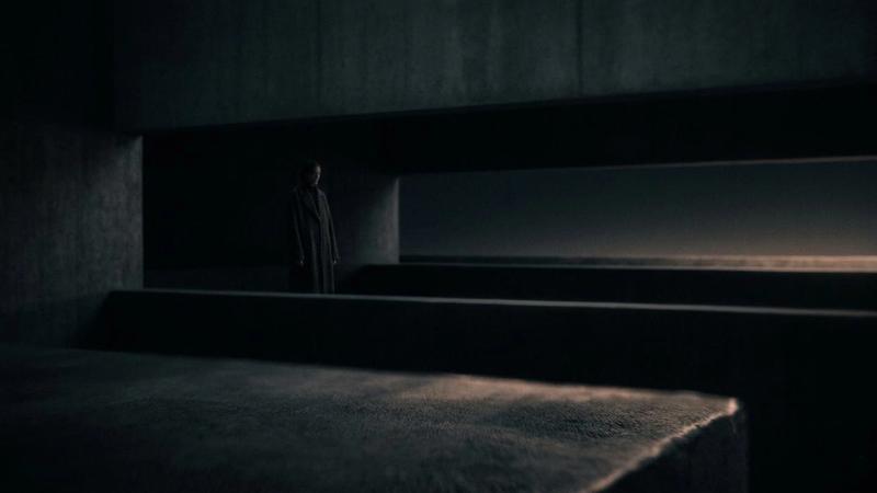 A cinematic, minimalist fine-art photograph in a 16:9 ratio with a dark, moody aesthetic. The composition features abstract, brutalist architectural layers in deep charcoal and midnight blue shadows. A soft, amber glow subtly catches the edge of a foreground slab, while a lone, quiet figure stands partially obscured in the distance between heavy horizontal planes. The image has a soft-focus lens effect and high-quality film grain, evoking a sense of restrained depth and structural mystery.