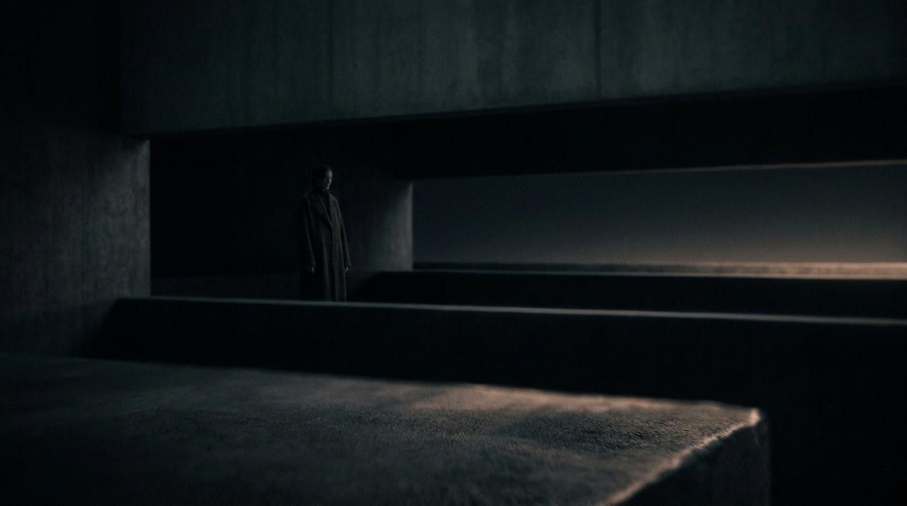 A cinematic, minimalist fine-art photograph in a 16:9 ratio with a dark, moody aesthetic. The composition features abstract, brutalist architectural layers in deep charcoal and midnight blue shadows. A soft, amber glow subtly catches the edge of a foreground slab, while a lone, quiet figure stands partially obscured in the distance between heavy horizontal planes. The image has a soft-focus lens effect and high-quality film grain, evoking a sense of restrained depth and structural mystery.