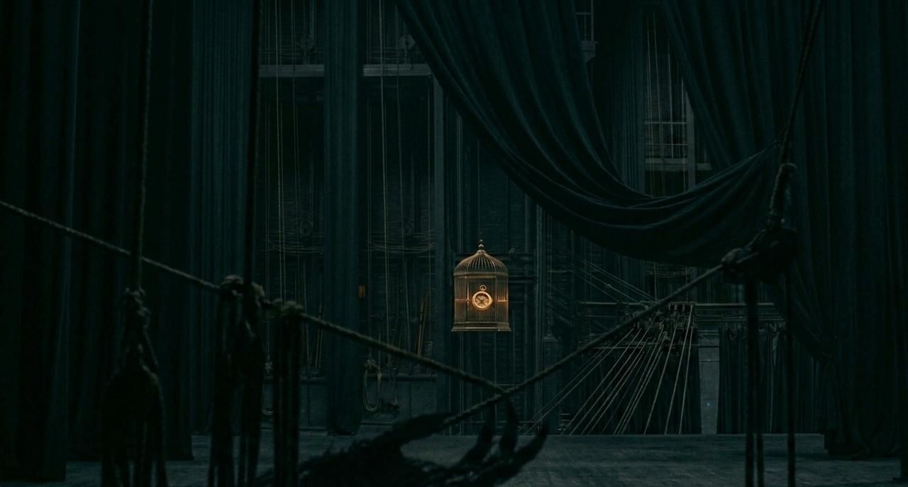 A dimly lit theater stage draped in dark curtains, with rigging ropes and a single illuminated birdcage hanging at the center, creating a dramatic and mysterious atmosphere.