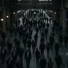 Blurred crowd moving through a dimly lit train station with arched architecture. Warm lights line the walls. Busy, hurried atmosphere.