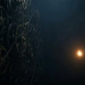 Dark cinematic scene featuring large metallic gears and machinery fading into shadows on the left side, with a small glowing spark of light floating in the empty dark space on the right, creating a mysterious and industrial atmosphere.