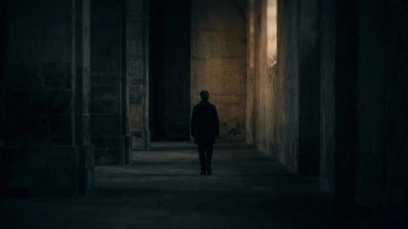 A person walks alone in a dim, narrow corridor with stone walls, creating a mysterious and somber mood. Light streams through a high window.