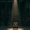 A single vintage armchair sits center stage under a narrow spotlight in a dark theater. Heavy black curtains frame the scene, while backstage rigging and ropes are faintly visible in the background, creating a dramatic and solitary atmosphere.