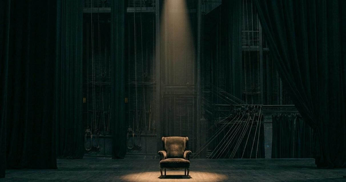A single vintage armchair sits center stage under a narrow spotlight in a dark theater. Heavy black curtains frame the scene, while backstage rigging and ropes are faintly visible in the background, creating a dramatic and solitary atmosphere.