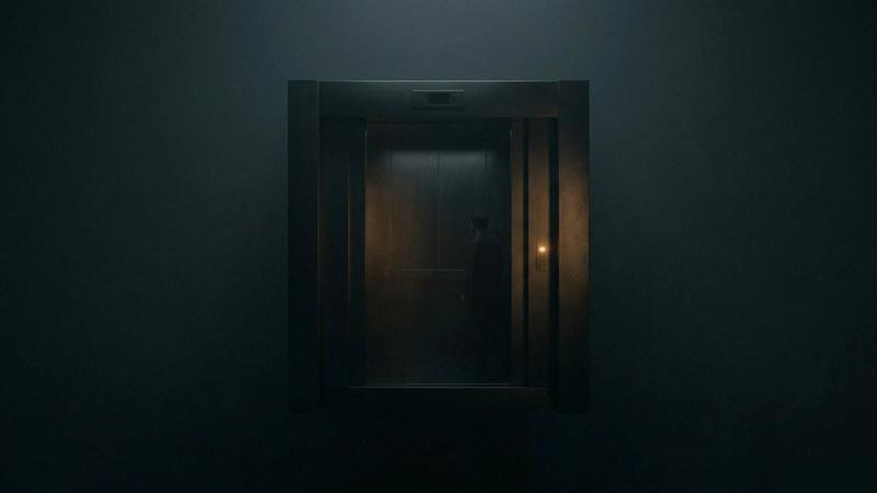 A man stands in a dimly lit elevator with closed doors. A subtle orange glow highlights muted gray walls, creating a mysterious atmosphere.