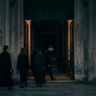 A dark, minimalist scene showing a glowing, arched doorway with a shadowy figure standing inside, partially reflected on a glossy floor, creating a mysterious and atmospheric mood.
