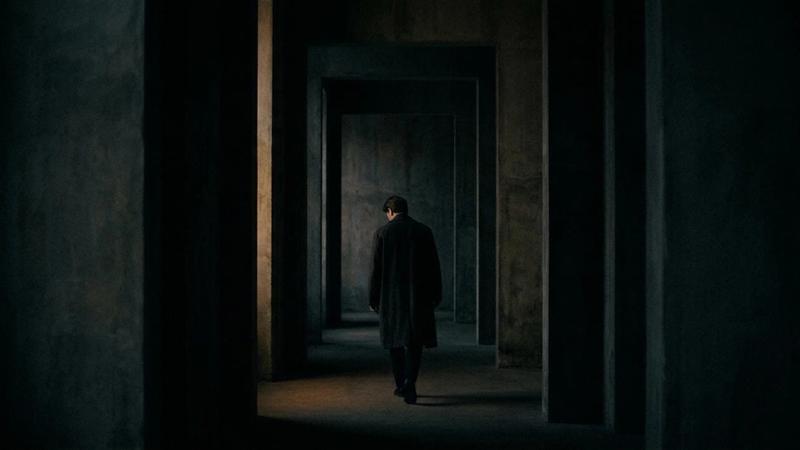 A solitary person in a dark coat walks through a dimly lit corridor of concrete walls, creating a somber and mysterious mood.
