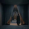 A dark, minimalist scene showing a glowing, arched doorway with a shadowy figure standing inside, partially reflected on a glossy floor, creating a mysterious and atmospheric mood.