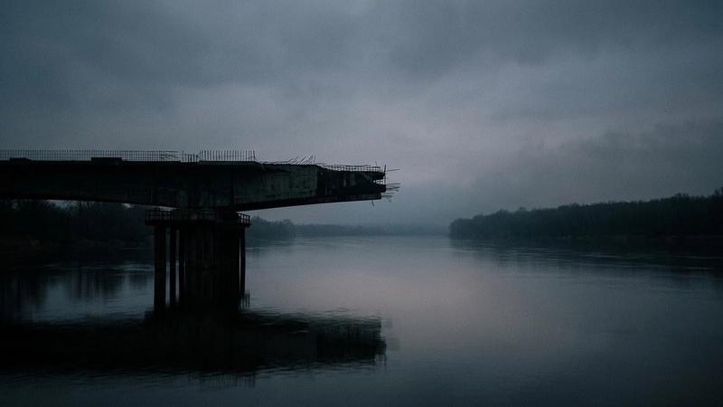 Broken bridge over a calm river under a cloudy, dim sky. The atmosphere is somber with dark water and silhouetted trees in the background.