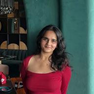 A woman wearing a red top stands indoors near a green pillar, smiling slightly, with a stylish restaurant setting and seating visible in the background.