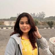 A woman wearing a yellow embroidered top and a gray hoodie stands outdoors near a roadside, gently touching her hair, with trees and a hazy sky in the background.