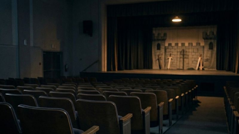 Empty auditorium with rows of seats facing a small stage set designed like a castle, illuminated by warm overhead lighting.