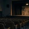 Empty auditorium with rows of seats facing a small stage set designed like a castle, illuminated by warm overhead lighting.