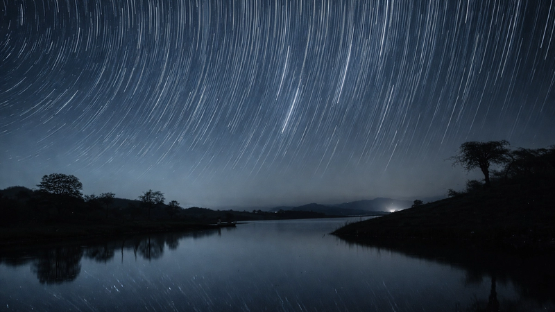Long-exposure star trails arc across a deep blue night sky above a calm lake, with dark silhouettes of trees and hills reflecting softly in the water.