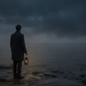 A wide, moody landscape photograph of a solitary man in a dark overcoat, viewed from behind, standing on rocky terrain at dusk. He holds a lit traditional lantern that provides the only warm light, looking out over a vast, fog-shrouded body of water under a dark, diffused sky.