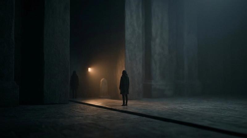 A solitary figure stands in a dim, shadowy hall with tall pillars. Soft light glows in the background, creating a mysterious, eerie mood.
