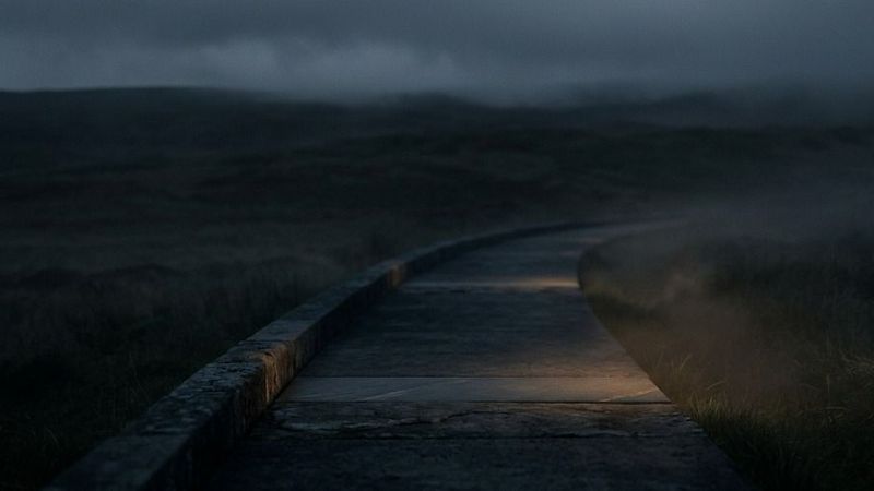 A dark, misty boardwalk winding through an open landscape under a cloudy evening sky, with low visibility and a moody atmosphere created by fog and dim light.