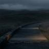 A dark, misty boardwalk winding through an open landscape under a cloudy evening sky, with low visibility and a moody atmosphere created by fog and dim light.