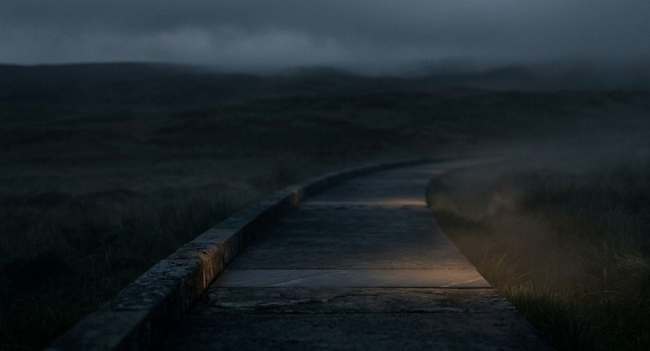 A dark, misty boardwalk winding through an open landscape under a cloudy evening sky, with low visibility and a moody atmosphere created by fog and dim light.