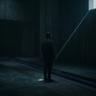 A solitary figure stands in a vast, dark concrete hall as a single beam of light cuts through the darkness, creating a cinematic and mysterious atmosphere.