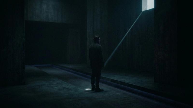 A solitary figure stands in a vast, dark concrete hall as a single beam of light cuts through the darkness, creating a cinematic and mysterious atmosphere.