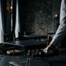 A minimalist and moody, wide-angle cinematic photograph of a high-end restaurant interior with a deeply desaturated palette of dark charcoals and midnight blues. In the foreground, a dimly lit dark wooden table holds two leather-bound menus. A sommelier, seen from the torso down in a crisp light shirt and a dark vest, subtly extends a hand to present a bottle of red wine over the table. The lighting is soft and diffused, creating heavy shadows and a quiet, melancholic editorial feel with high-quality film grain and a soft-focus lens.